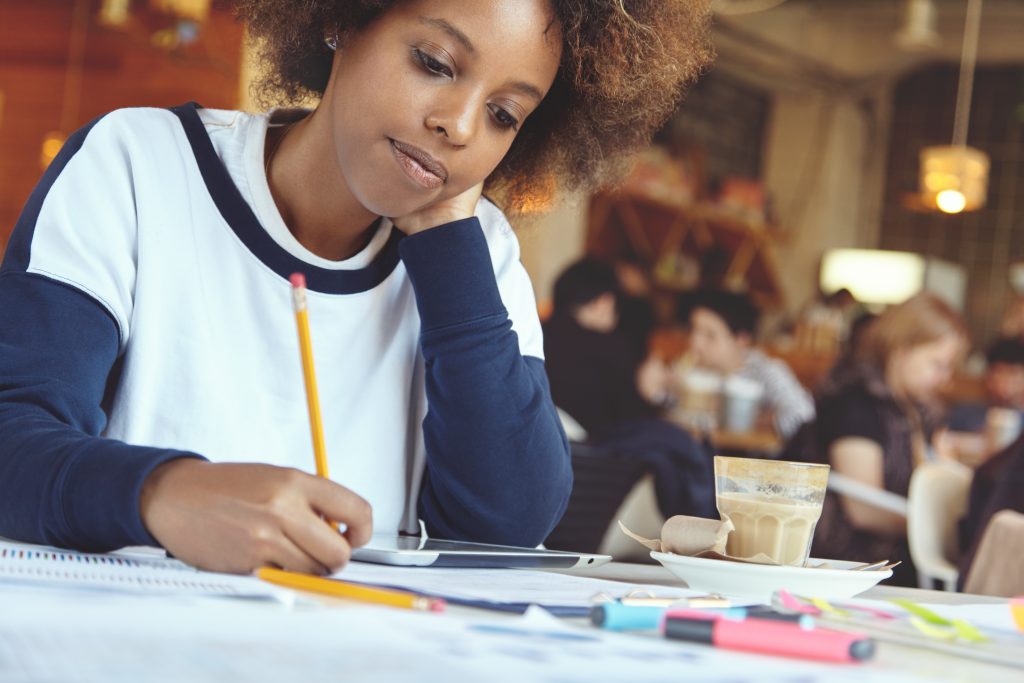headshot of beautiful young african female freelancer writing down plans on her project using touch pad at coffee shop, making notes with pencil, sitting at table with papers, drinking hot chocolate